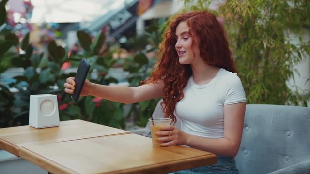Beautiful young curly ginger blogger girl having an online streaming chat with her followers while being in a café, cutely smiling, showing coffee cup. Modern communication, technologies concept