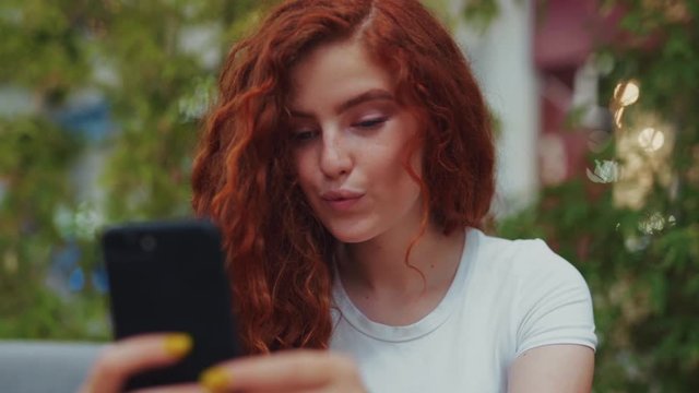 Close up view of lovely curly ginger student girl sitting in café alone and actively chatting with her friends, followers via smartphone. Online, browsing social networks, communication and