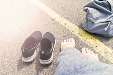 Fototapeta premium Woman with bare feet and backpack sitting on an asphalt road with a yellow single road line waiting vehicle for hitchhiking, summer season