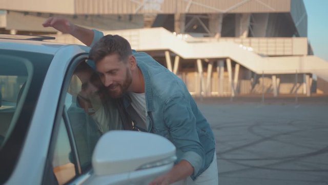 Cheeful handsome man leaning hugging the side of new modern car enjoying a successful purchase at dealership on parking lot.