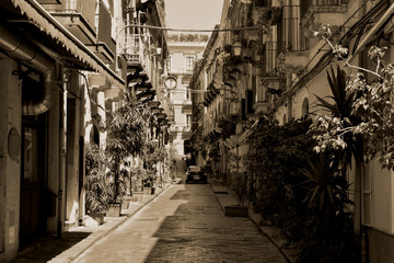 Typical Italian street in Catania. Narrow paved road with little trees and houses with small balconies on both sides. Black and white
