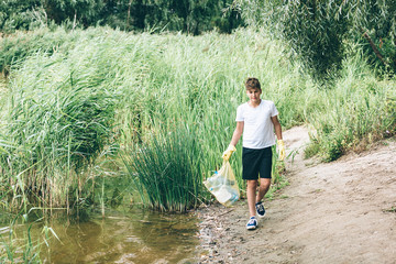 Boy in white t shirt in gloves collects garbage and plastic bottles into blue package on the beach....