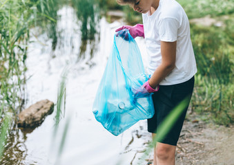 Boy in white t shirt in gloves collects garbage and plastic bottles into blue package on the beach. Young volunteer. Environmental protection, save environment concept