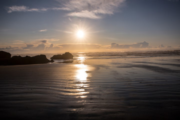 Naklejka premium Cannon Beach w stanie Oregon. Sunburst, gdy słońce zachodzi nad Oceanem Spokojnym, baseny pływowe, skały stogu siana i mokry piasek