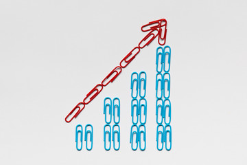 A red arrow pointing upwards siting on a stairwell made from blue paperclips on white background, shot from above, closeup.