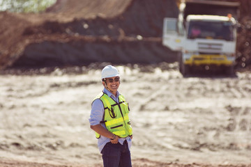 portrait mining worker or engineer in reflective vest and hardhat posing with hand inside pocket in front of white truck at coal and soil mining site