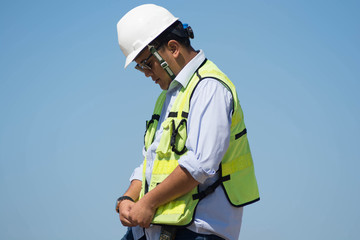 bottom rear view of male engineer walking and holding tablet computer on bright blue sky for planing a construction dress with protective vest, hard helmet
