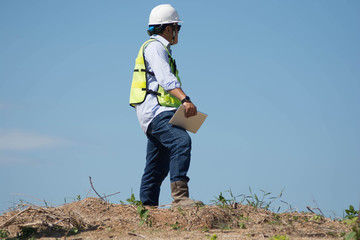 bottom rear view of male engineer walking and holding tablet computer on bright blue sky for planing a construction dress with protective vest, hard helmet