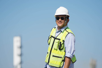 Portrait of Confident Asian engineer. Handsome young man in hardhat using glasses while standing outdoors and against blue sky