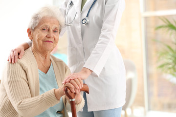 Nurse assisting elderly woman with cane indoors. Space for text