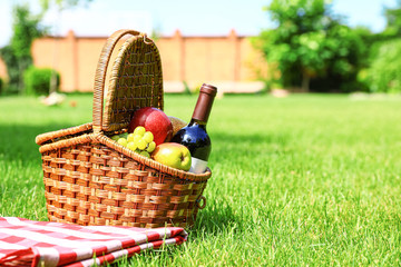 Picnic basket with fruits, bottle of wine and checkered blanket on green grass in garden. Space for text