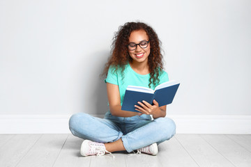 Fototapeta premium Beautiful young African-American woman reading book on wooden floor near light wall