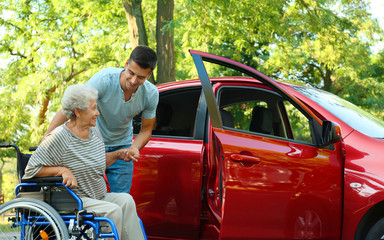 Young man helping disabled senior woman in wheelchair to get into car outdoors