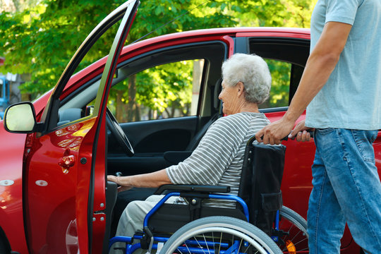 Young man helping disabled senior woman in wheelchair to get into car outdoors