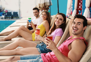 Happy young friends with fresh summer cocktails relaxing on sunbeds near swimming pool
