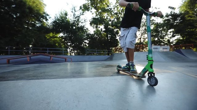 boy in t-shirt and shorts rides scooter with handlebar on grey funbox with tricks against green trees slow motion