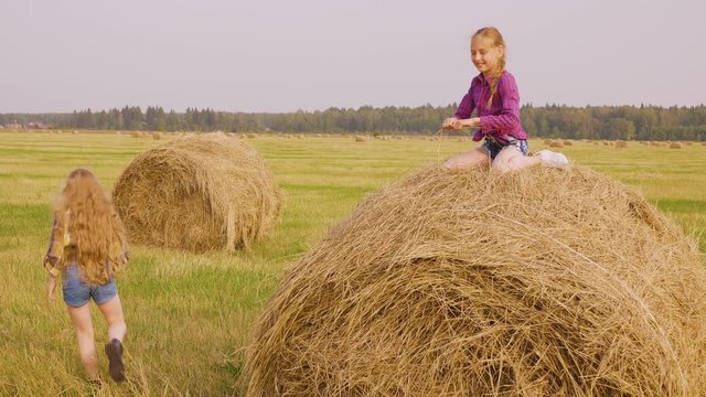 Summer vacation on countryside, village, farm. Outdoor nature activities of teen girls. Girl is trying to climb on hay stack to her friend sitting on top and helping her holding hand and pulling.