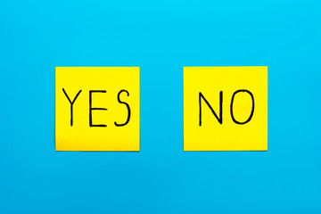 Close up black handwritten inscription yes and no word on two yellow square stickers on blue background with copy spase, horizontal orientation