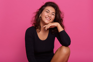 Close up portrait of attractive young woman keeping hand under chin, smiling while sitting against pink background, wearing black shirt, being in good mood, having dark wavy hair and stylish earrings.