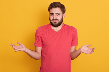 Doubtful male isolated over yellow studio background, unsure man keep hands aside doubt over deal, make decision or choice, do not know what to choose, wearing red casual t shirt. People emotions.