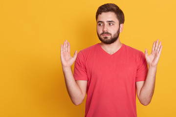 Studio shot of serious casual man keeps hands up isolated over yellow background, handsome male looking aside, having beard, wearing red t shirt. Copy space for your advertisment or promotion text.