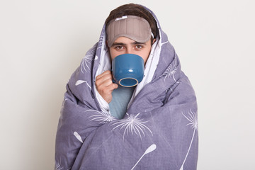 Studio shot of unhealthy male wrapped in wool blanket holding blue mug while being indoors, drinking herbal tea, has headache and catches cold, posing with blindfold on forehead, isolated over white.