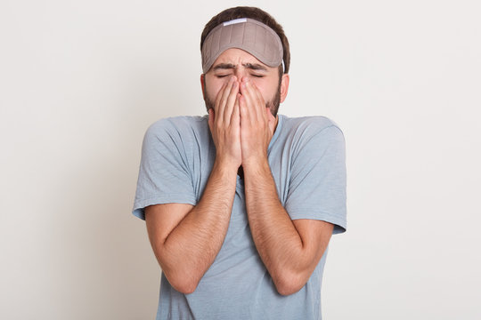 Image of young man with beard wearing casual gray t shirt over white isolated background, unshaven man yawning, looks tired, covering his face with hands, posing with closed eyes, wants to sleep.