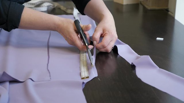 Cutting and sewing products in tailoring business. Tailor woman cutting fabric using large scissors following chalk markings of pattern, closeup hands. Dressmaker creating new clothes.