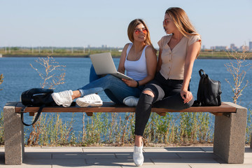 Two young female girlfriends having fun while sitting on a bench while one showing to screen of a laptop while another one is laughing outside.