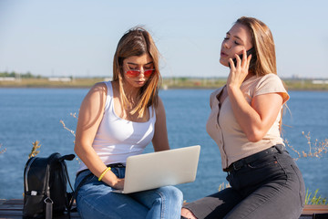 Fototapeta premium Two smiling young girls using laptop at the park, laughing, chatting