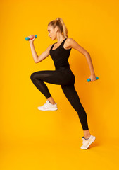 Young Woman Exercising Jumping With Dumbbells On Yellow Studio Background