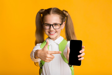 Fototapeta premium Smiling Schoolgirl Pointing Finger At Blank Cellphone Screen, Yellow Background