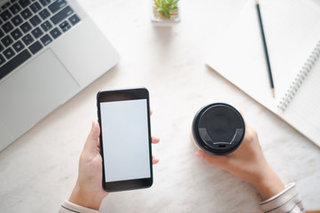 Closed up and focus on hand of young woman using a smartphone which show white display other hand hold a cup of coffee and have notebook put on table in near. see top view. co working space concept.