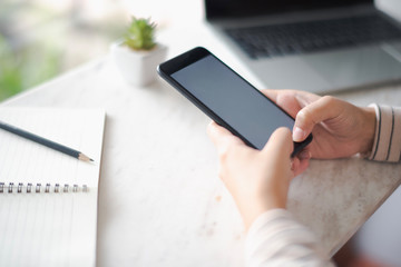 Closed up and focus on hand of young woman using a smartphone which show white display and have notebook put on table near. co working space concept.