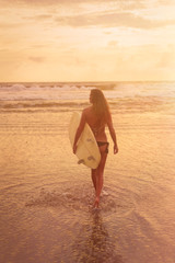 A young woman in bikini stands with her back to the camera and holds a surfboard by the ocean. Warm tinting