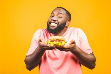 Young african american man eating hamburger isolated over yellow background.
