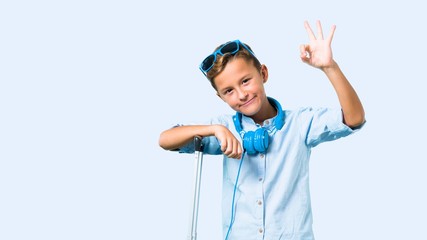Kid with sunglasses and headphones traveling with his suitcase showing an ok sign with fingers on blue background