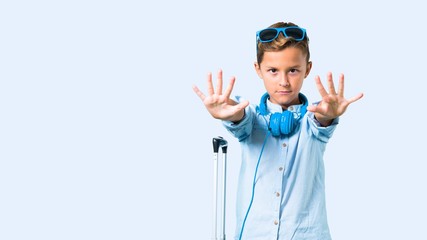Fototapeta premium Kid with sunglasses and headphones traveling with his suitcase making stop gesture with her hand on blue background