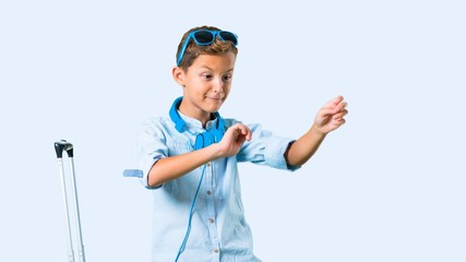 Kid with sunglasses and headphones traveling with his suitcase listening to the music and dancing on blue background