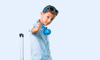 Kid with sunglasses and headphones traveling with his suitcase handshaking after good deal on blue background