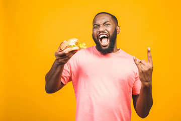 Young african american man eating hamburger isolated over yellow background.