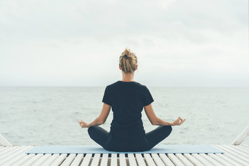 A young woman in a yoga asana with her back to the camera meditates while looking at the sea waves. Morning exercise in solitude and peace.