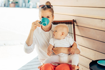 Caucasian young woman sits in a deck chair at the table, drinks coffee and holds the baby in her...