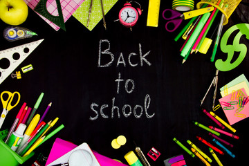 Chalk inscription of back to school on blackboard background. Pink, yellow and green office supplies, pens, rulers, paper clips, alarm clock are scattered around of black canvas. Preparing for study