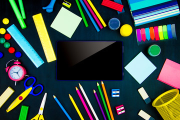 Back to school, college, university. Blue tablet computer in center of blackboard background. Office supplies, pencils, markers, scissors, sharpeners, rulers, alarm clock are scattered on canvas.