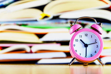 Pink alarm clock on background of stack of open colorful books on classroom desk. Preparing pupils...