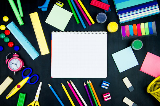 Back to school, college, university. White page of notebook in center of blackboard background. Office supplies, pencils, scissors, sharpeners, plasticine, alarm clock are scattered on black canvas.