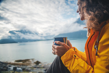 A young beautiful girl is drinking tea on the shore of the fjord, a trip to Norway, the Nordic...