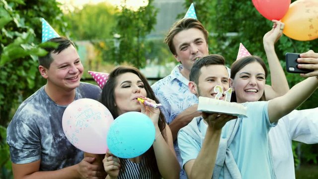Happy man makes selfie on mobile phone while celebrates a birthday, friends congratulate with balloons, cake and party whistle, take a photo on mobile phone
