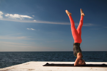 Adult blond woman with short haircut practices yoga on the pier against the background of the sea and blue sky